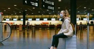 A Woman Wearing A Face Mask Sits On Luggage In An Airport Terminal Amid The Pandemic.