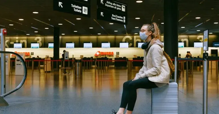A Woman Wearing A Face Mask Sits On Luggage In An Airport Terminal Amid The Pandemic.