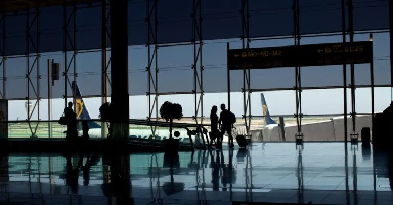 Travelers silhouetted in a modern airport terminal with glass windows and visible airplane tails.