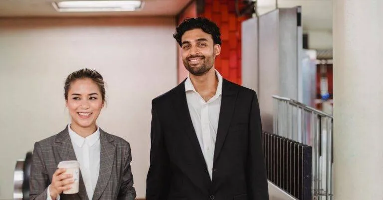 Two Smiling Professionals Walking In A New York Subway Station, Holding Coffee And Briefcase.