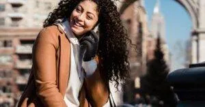 Young Woman With Curly Hair Happily Chatting On Phone In A City Setting, Enjoying A Sunny Day.