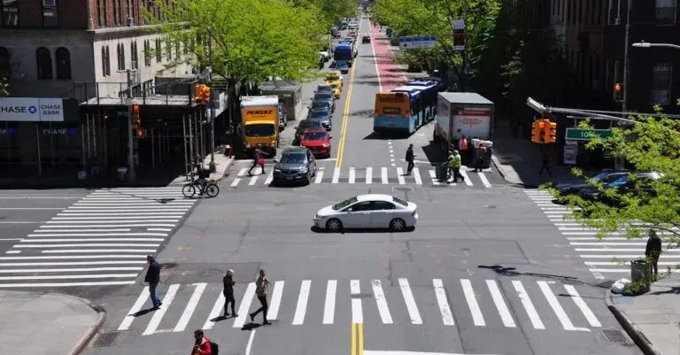A Bustling New York City Intersection With Cars, Buses, And Pedestrians On A Sunny Day.
