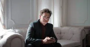 Businessman In A Black Suit Sitting On A Sofa, Using A Smartphone. Bright And Modern Indoors Setting.