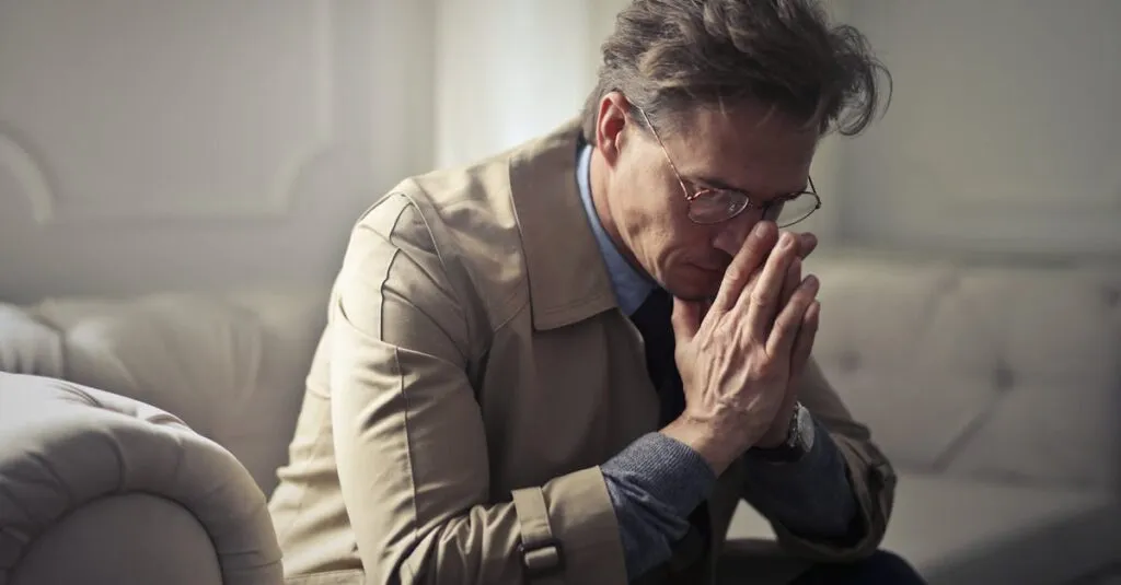 Side view of upset male entrepreneur in formal wear sitting on couch in modern living room and contemplating about financial problems while leaning on hands and looking down
