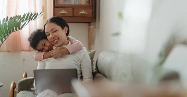 Excited Cute Asian Daughter Bonding With Smiling Happy Mother Using Netbook On Cozy Sofa In Living Room In Daytime