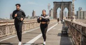 Two Adults Jogging On Brooklyn Bridge With City Skyline In The Background, Showcasing Health And Vitality.