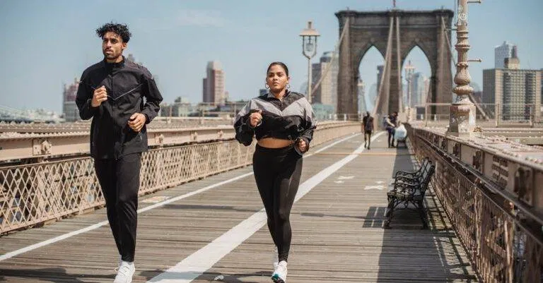 Two Adults Jogging On Brooklyn Bridge With City Skyline In The Background, Showcasing Health And Vitality.