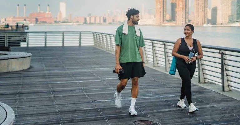 A Man And Woman In Sportswear Walking And Exercising On A Riverside Boardwalk With A Cityscape View.