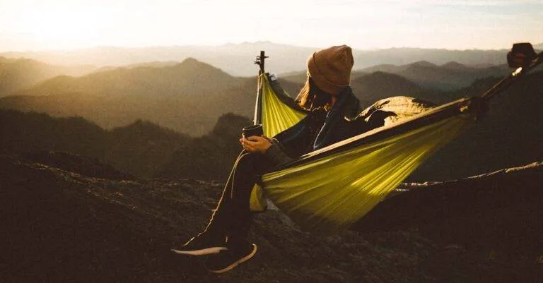 Woman Relaxing In A Hammock With A Hot Drink Amidst Majestic Mountain Scenery At Sunset.