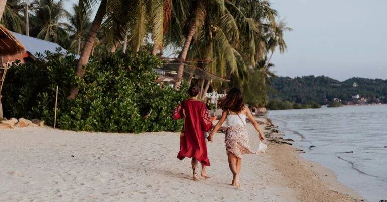 Two Women Holding Hands And Walking Along A Sandy Tropical Beach, Enjoying Their Summer Vacation.