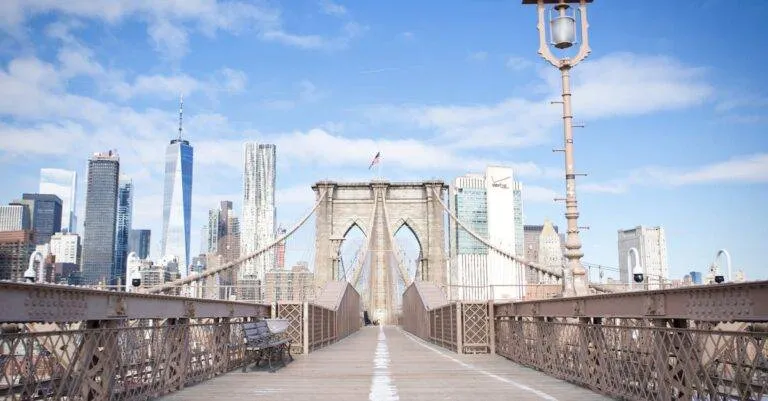 Wide-Angle View Of The Brooklyn Bridge Leading To New York City'S Iconic Skyline Under Blue Skies.