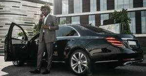 Elegantly Dressed Man In Gray Suit Making Phone Call Next To Sleek Black Car.