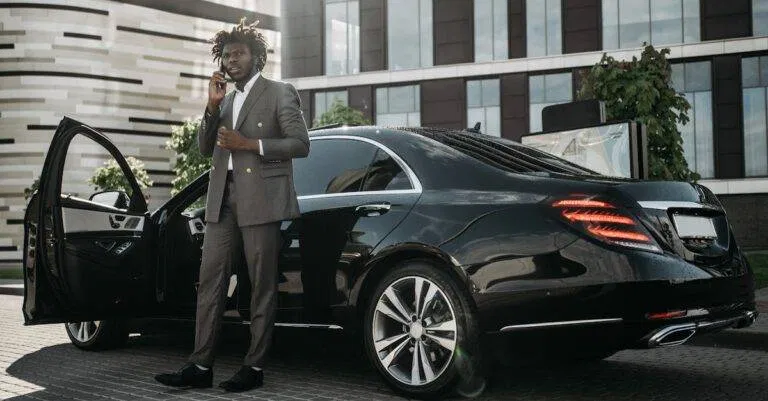 Elegantly Dressed Man In Gray Suit Making Phone Call Next To Sleek Black Car.