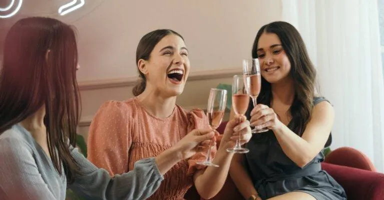 Three Women Laughing And Clinking Champagne Glasses In A Joyful Indoor Celebration.