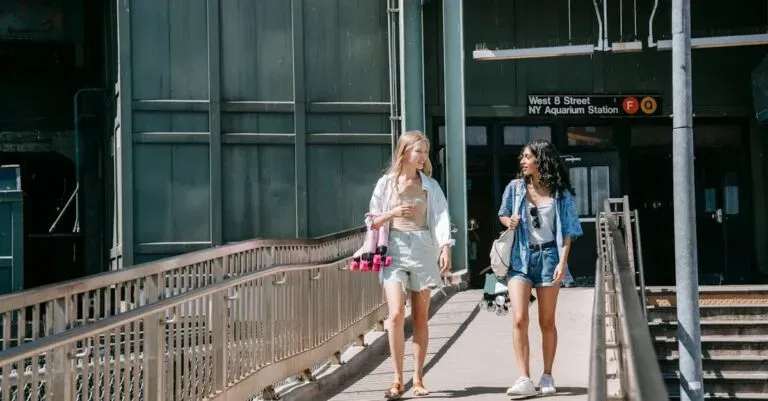 Two Young Women Casually Walking On A Footbridge Outside Ny Aquarium Station On A Sunny Day.