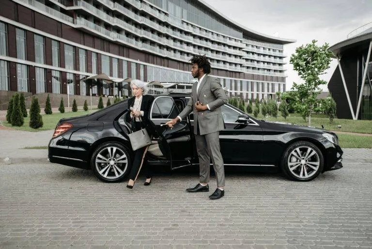 A Professional Chauffeur Assists A Businesswoman Exiting A Luxury Car In Front Of A Modern Building.