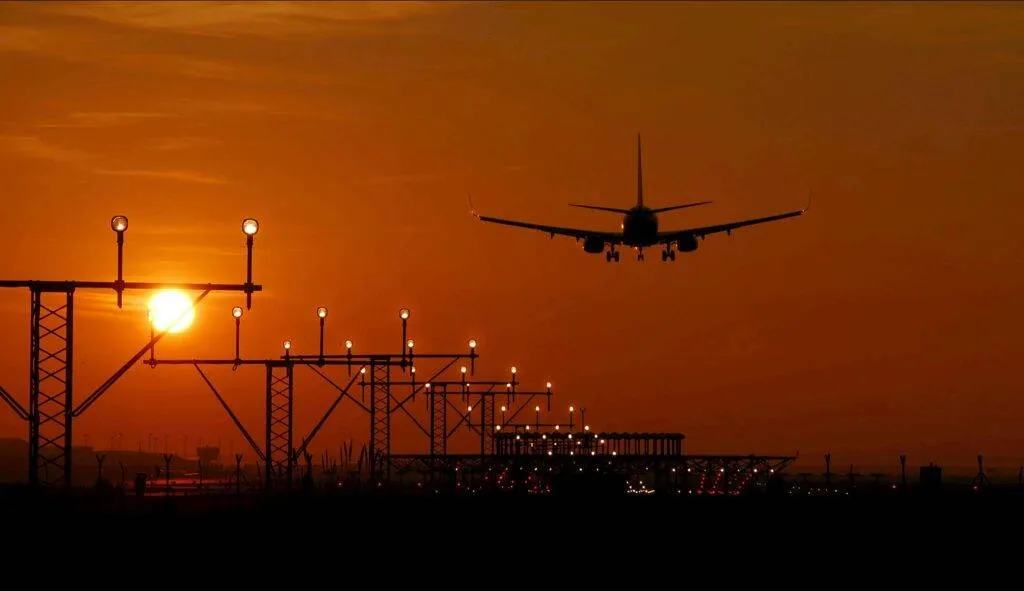 Silhouette Of An Airplane Landing During Sunset At Barcelona Airport, With A Vibrant Orange Sky.