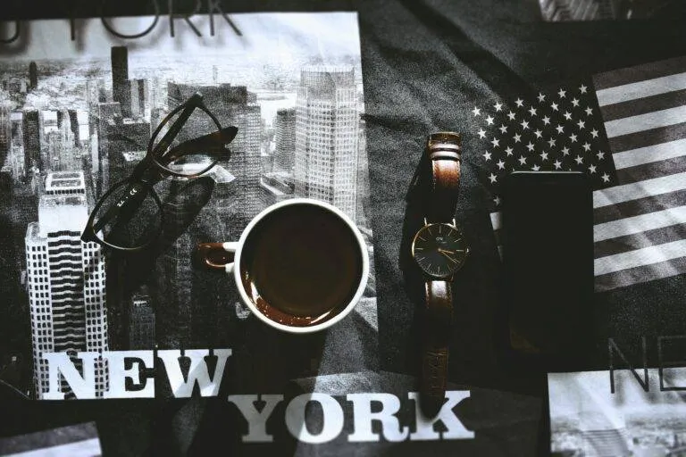 Stylish Flatlay Showcasing A Cup Of Coffee, Wristwatch, Eyewear, And Phone On A New York Themed Background.