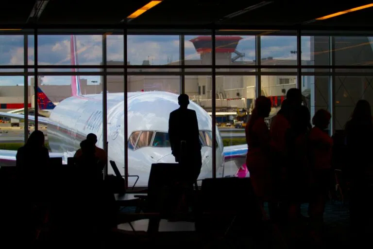 Silhouettes Of Travelers Waiting At An Airport Terminal With An Airplane Visible Through The Window.