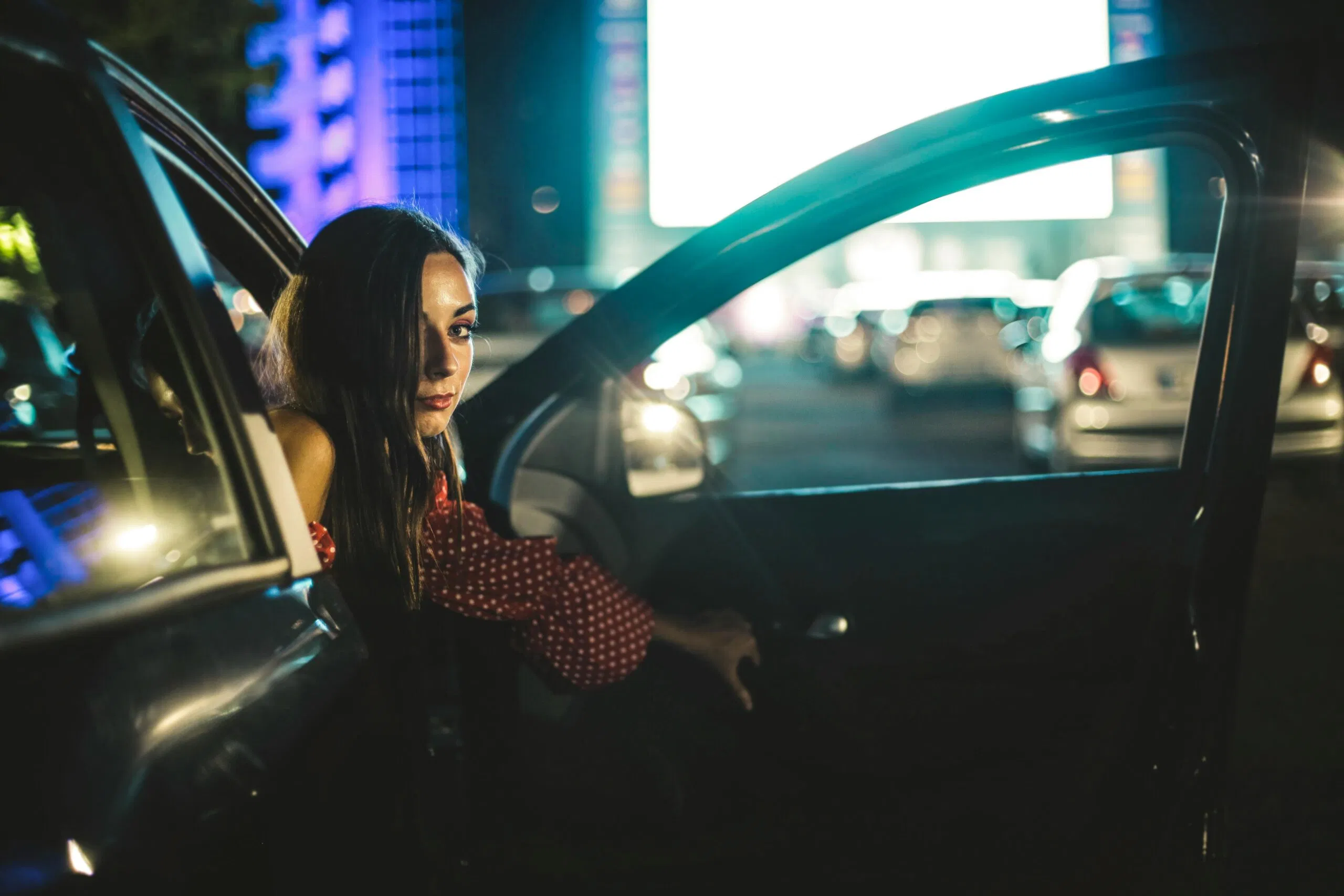 Attractive Female With Long Hair Covering Half Face Looking At Camera While Sitting In Car With Opened Door On Parking Lot Against Blurred Screen