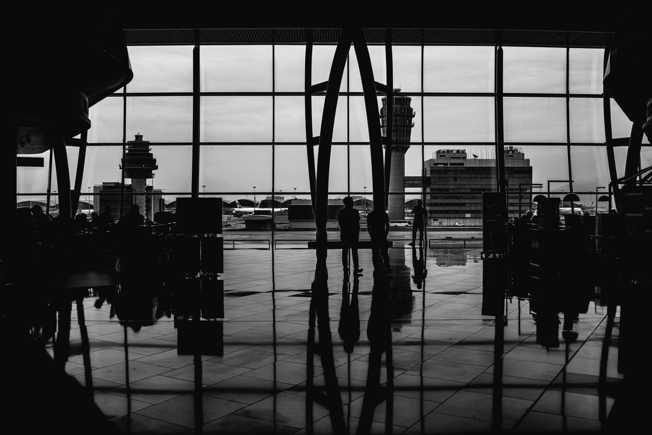 Lga And Jfk Distance: 7 Honest Facts Families Need In 2026 2 August 23, 2026 Black And White Image Of Travelers Silhouetted Against Large Airport Windows, Capturing Urban Travel Ambiance.